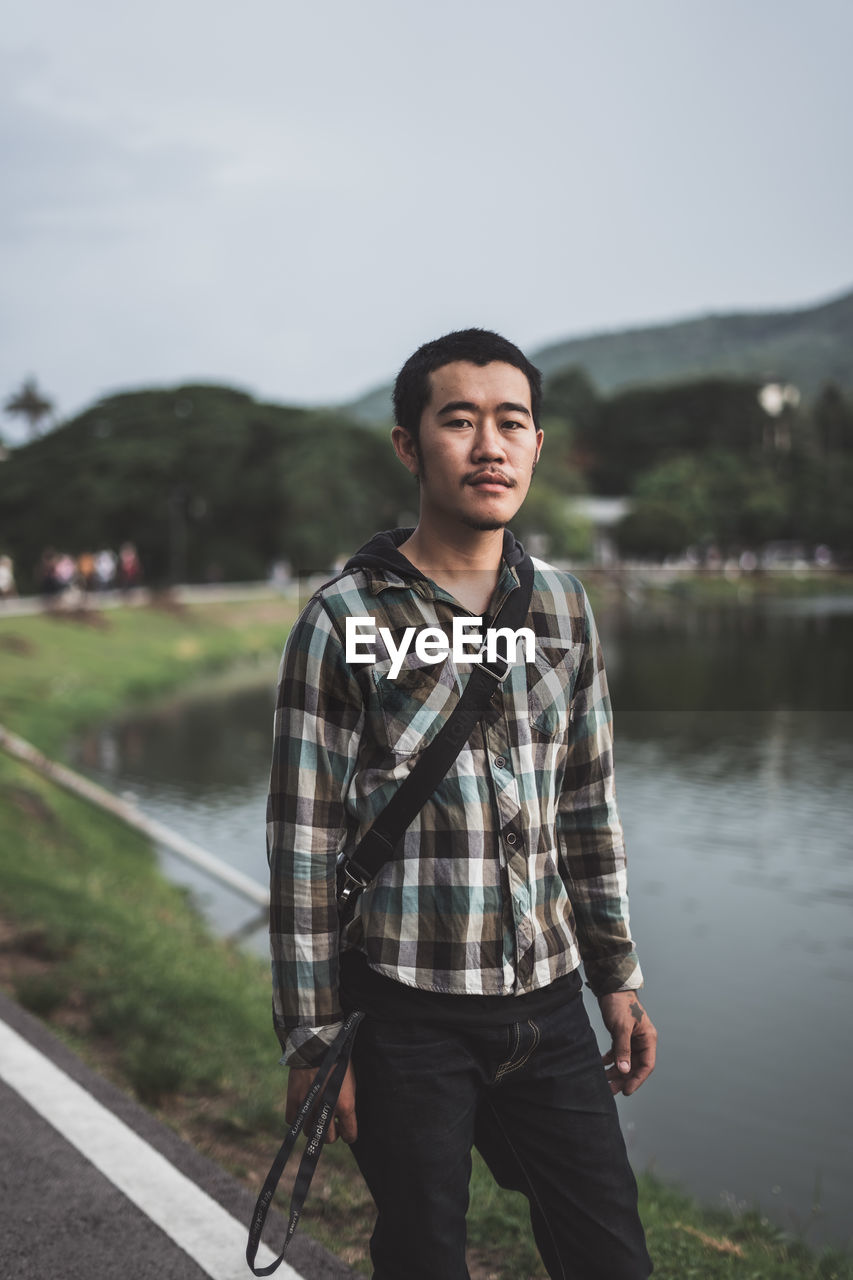 PORTRAIT OF A YOUNG MAN STANDING AGAINST LAKE