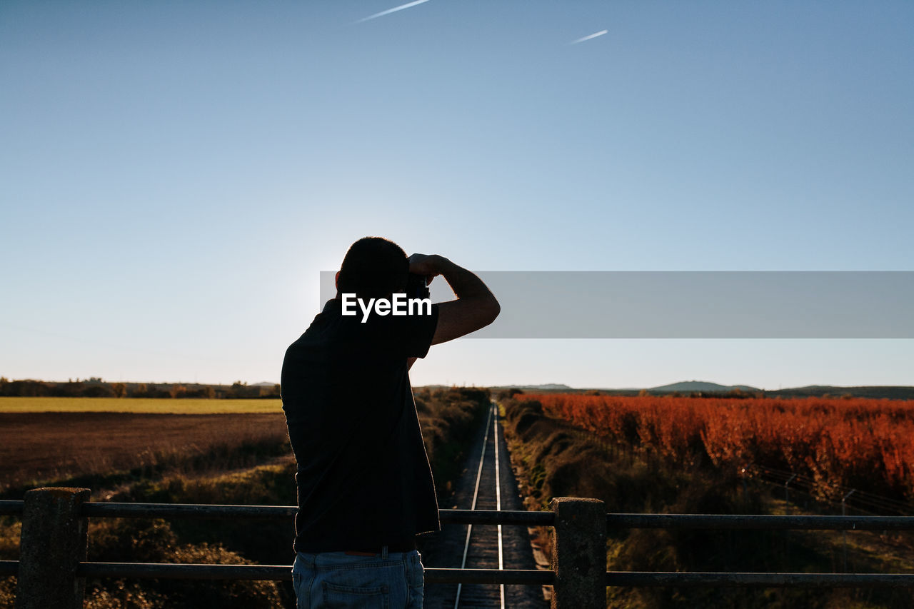 Rear view of man photographing railroad tracks from bridge against sky