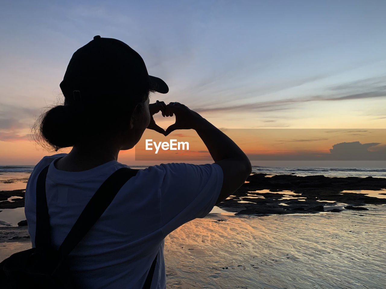 Silhouette of a young woman making heart love sign against colorful sunset on the beach. 