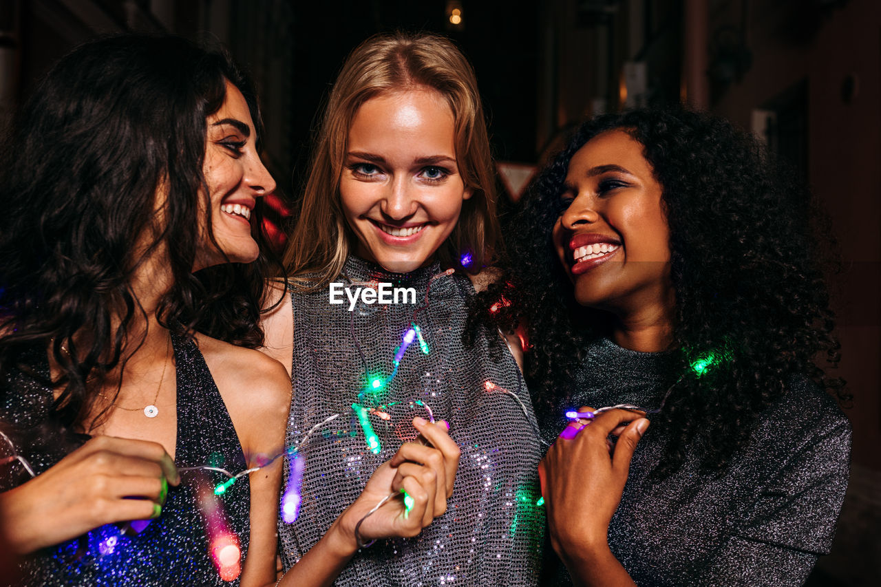 Cheerful female friends holding string light outdoors at night