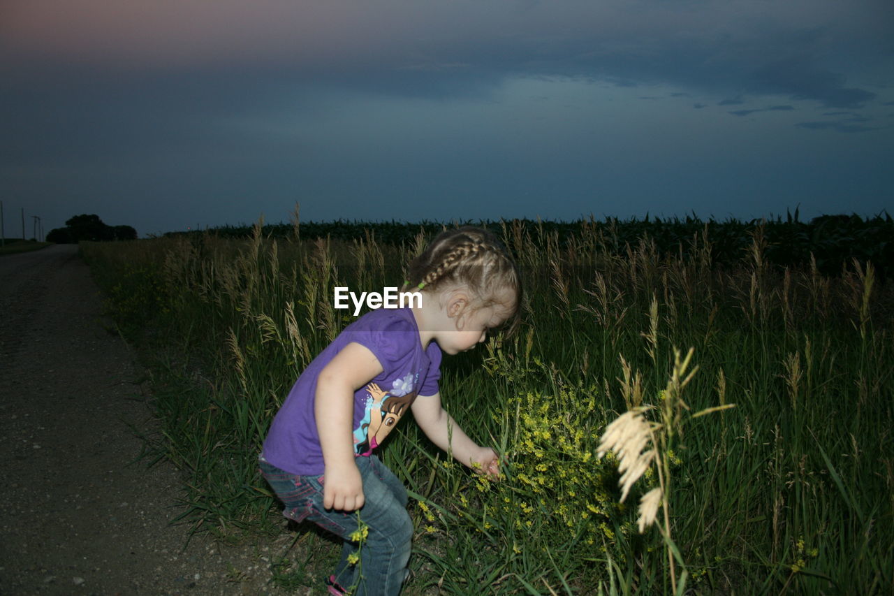 Side view of girl touching flowers at roadside against sky