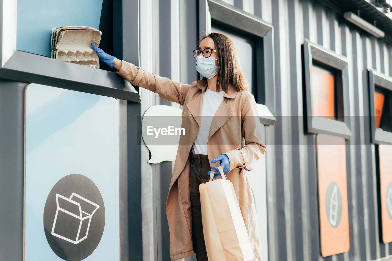Young woman in protective face mask and rubber gloves throwing garbage into recyclable trash.