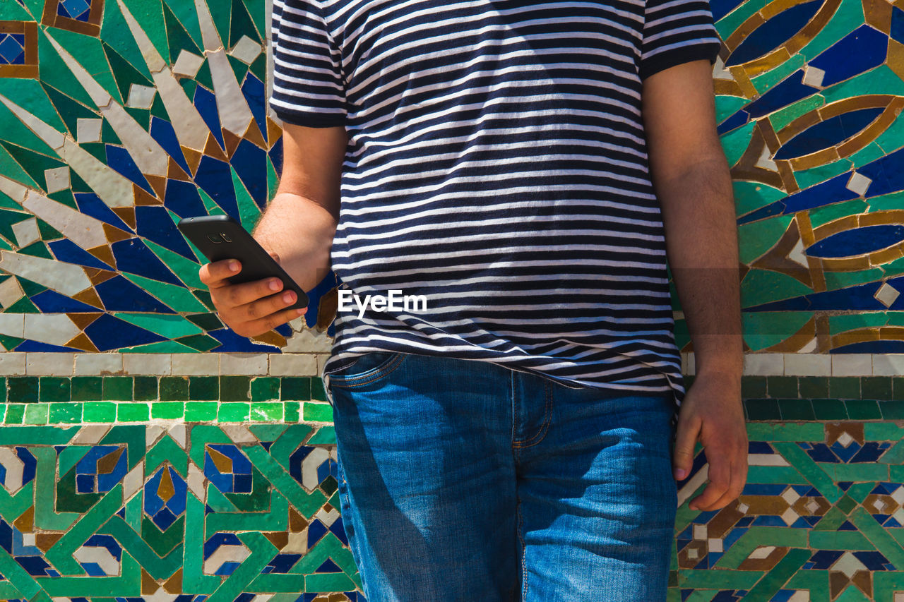 Midsection of man using mobile phone while standing against patterned wall 
