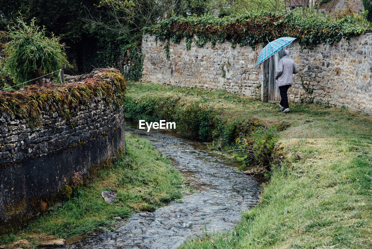 Rear view full length of senior man with umbrella walking by stream on grass