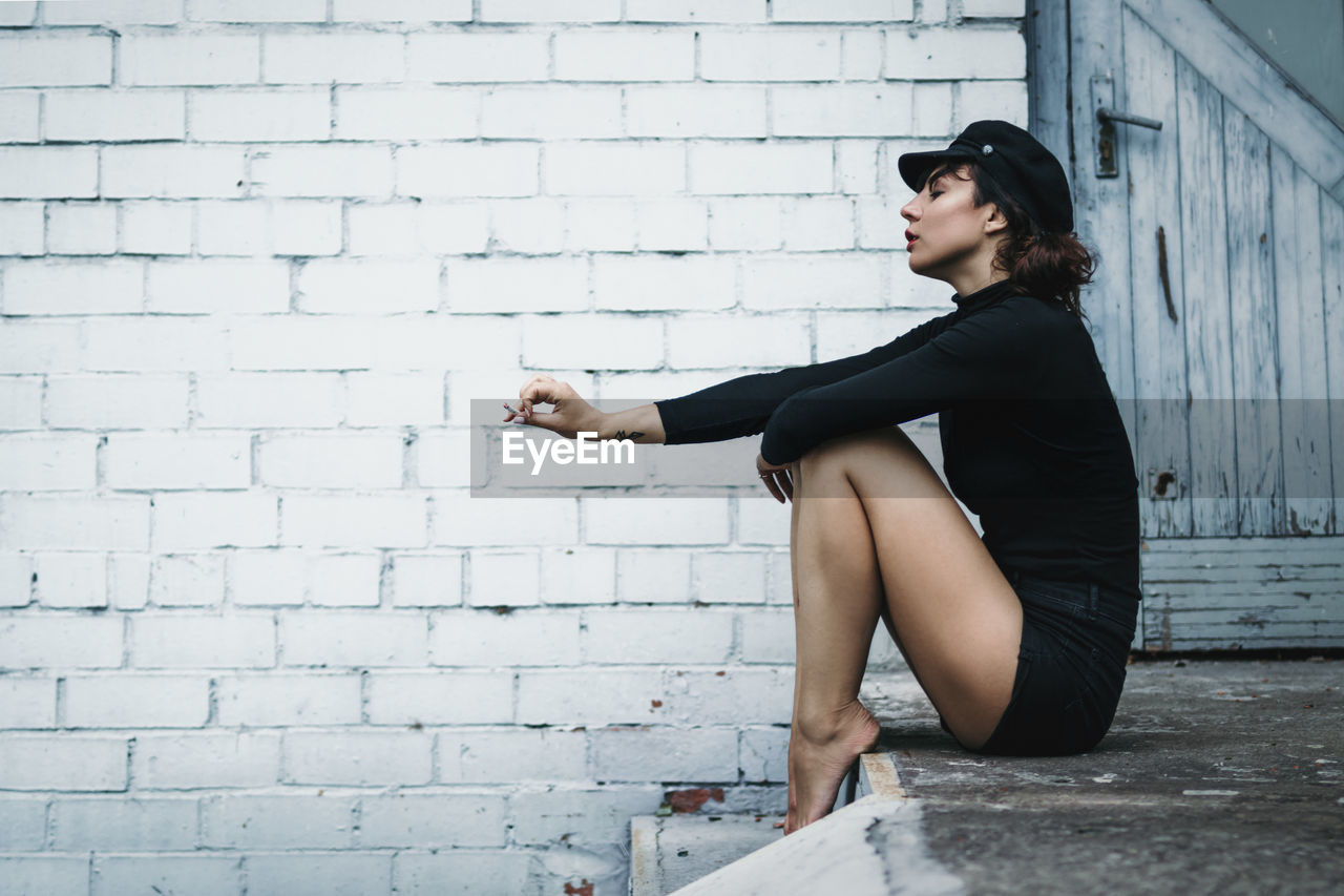 Side view of mid adult woman smoking while sitting against brick wall