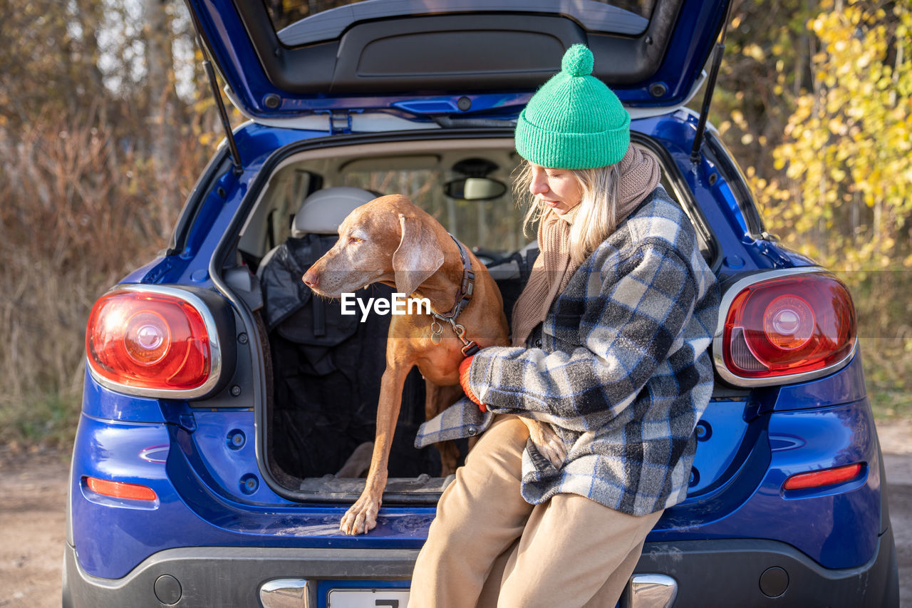 Pet owner woman in road trip holding dog on leash sitting in car trunk on nature.