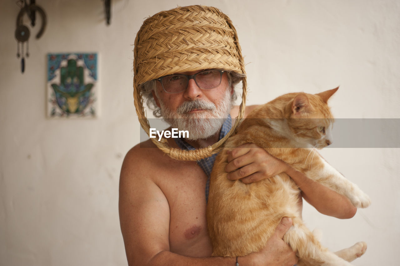 Portrait of shirtless senior man with basket on head holding cat against wall at home