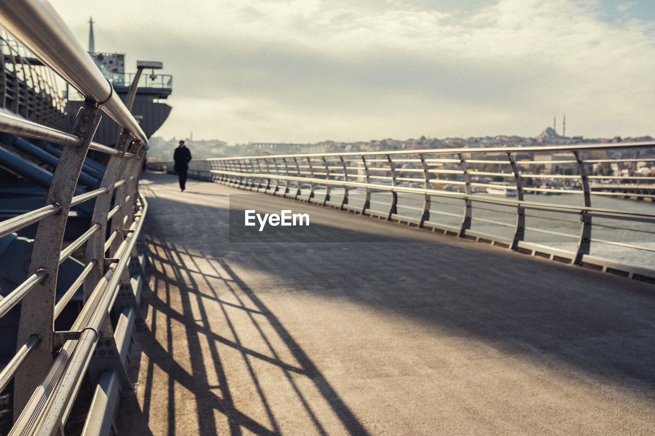 REAR VIEW OF MAN WALKING ON BRIDGE ROAD AGAINST CLOUDY SKY