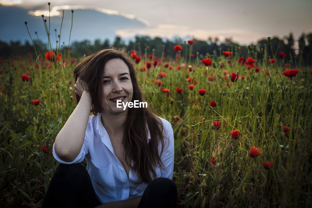 Portrait of smiling young woman sitting by poppy flowers on field