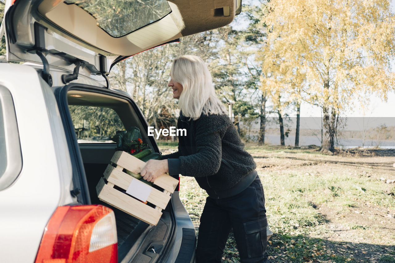 Side view of mature woman loading crate full of organic vegetables in car trunk