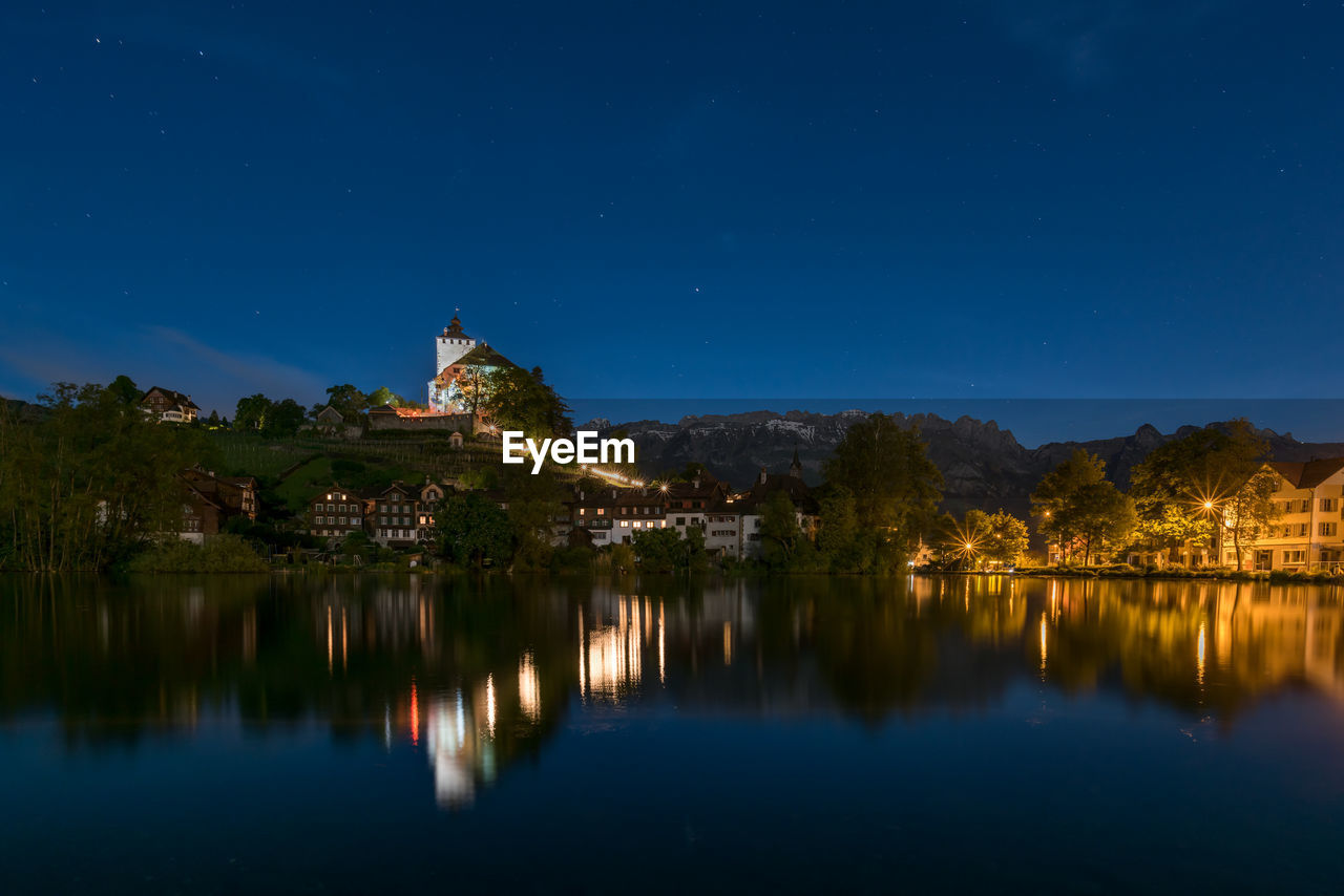 Reflection of buildings on calm lake against blue sky at night