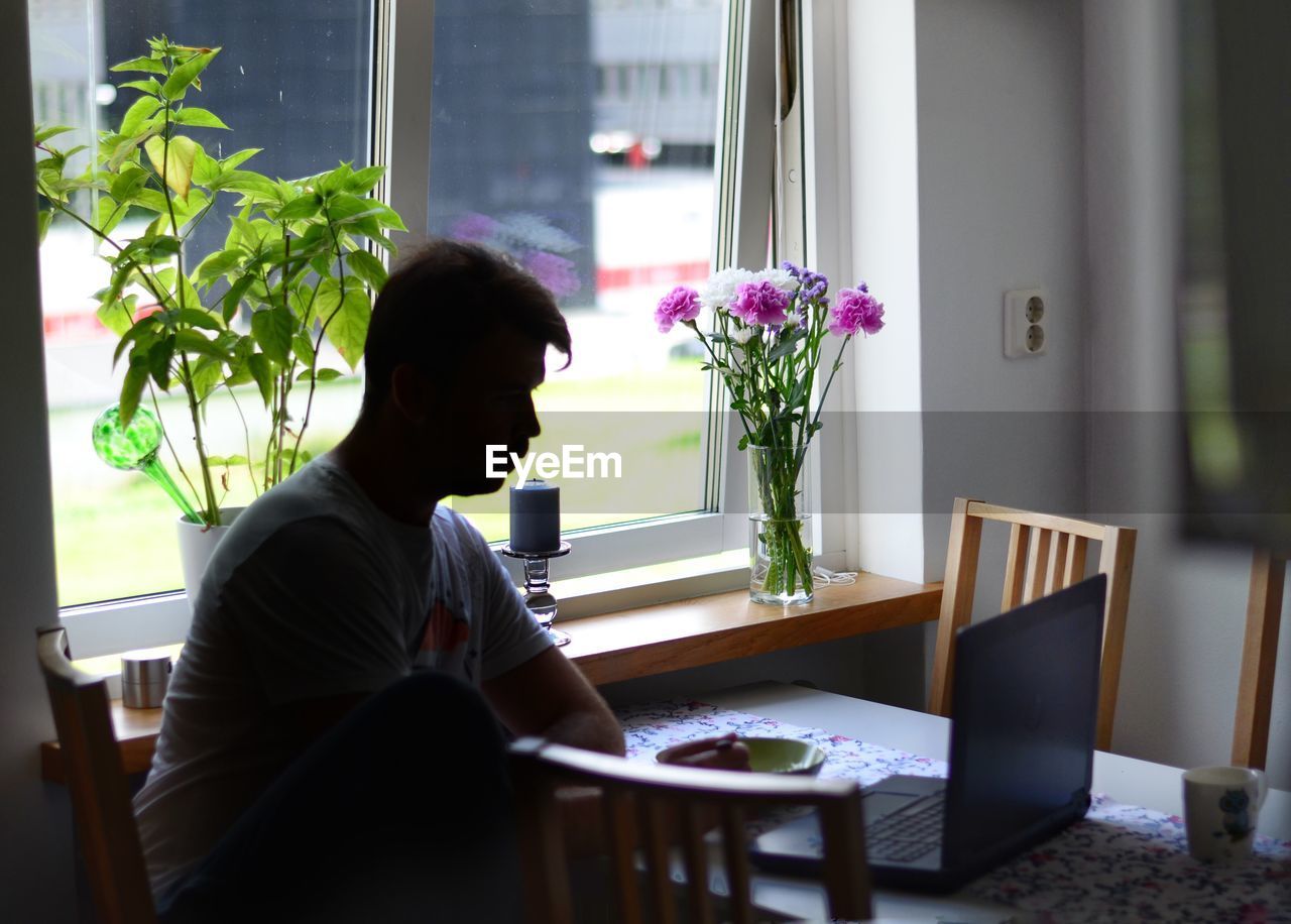 Silhouette man using laptop at home while sitting on chair near window