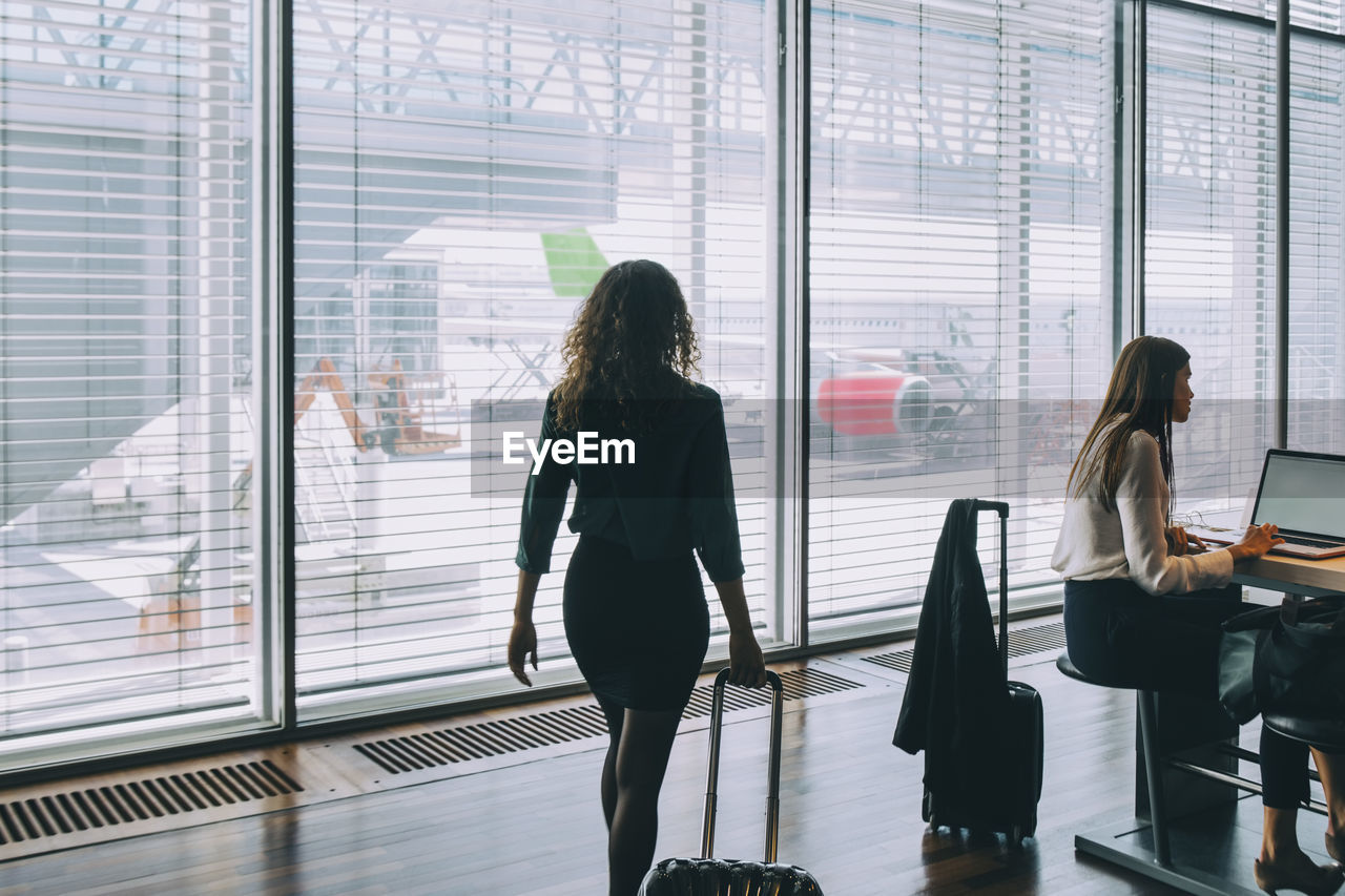 Rear view of businesswoman walking with luggage by female colleague sitting at airport departure area