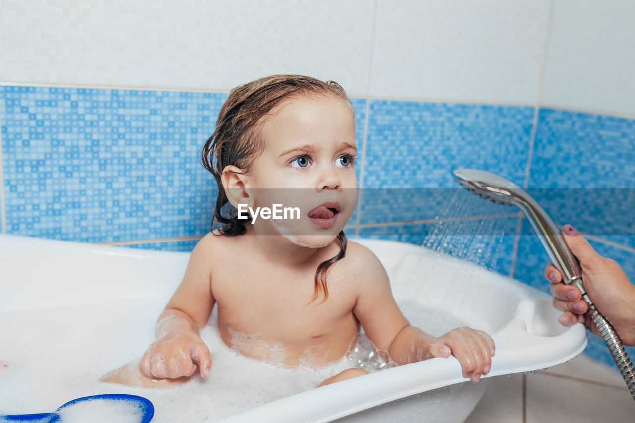 Cropped hand of mother showering shirtless daughter sitting in bathtub