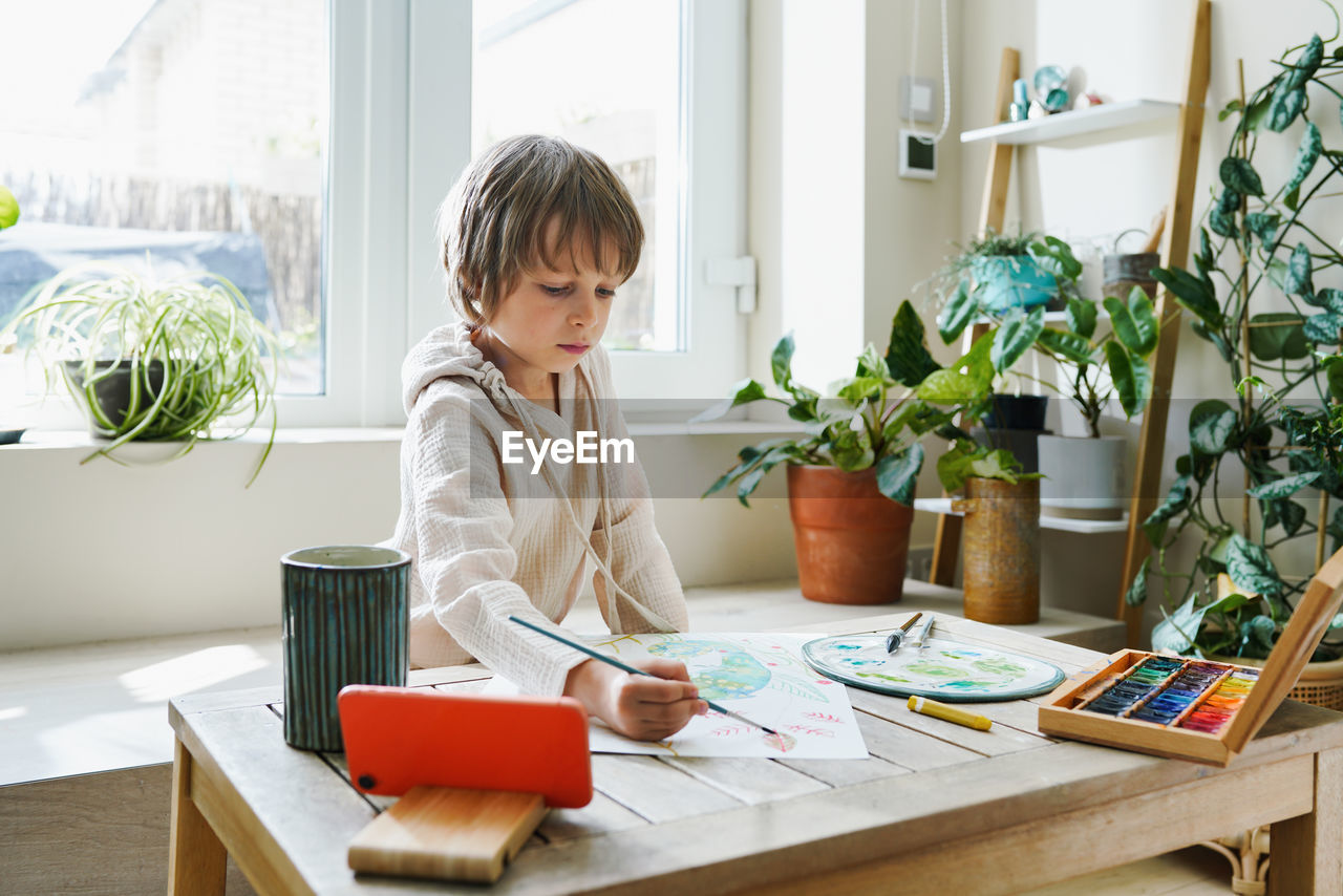 Cute male kid art drawing multicolored image use paints and brush on table at brightly room