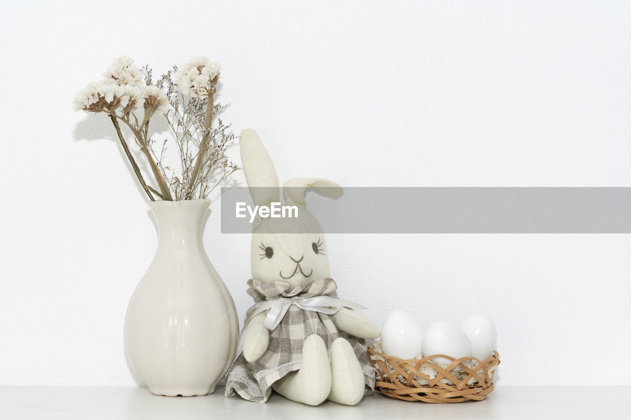 Easter composition  soft rabbit igushka, white eggs in a basket and a vase of flowers on a shelf 