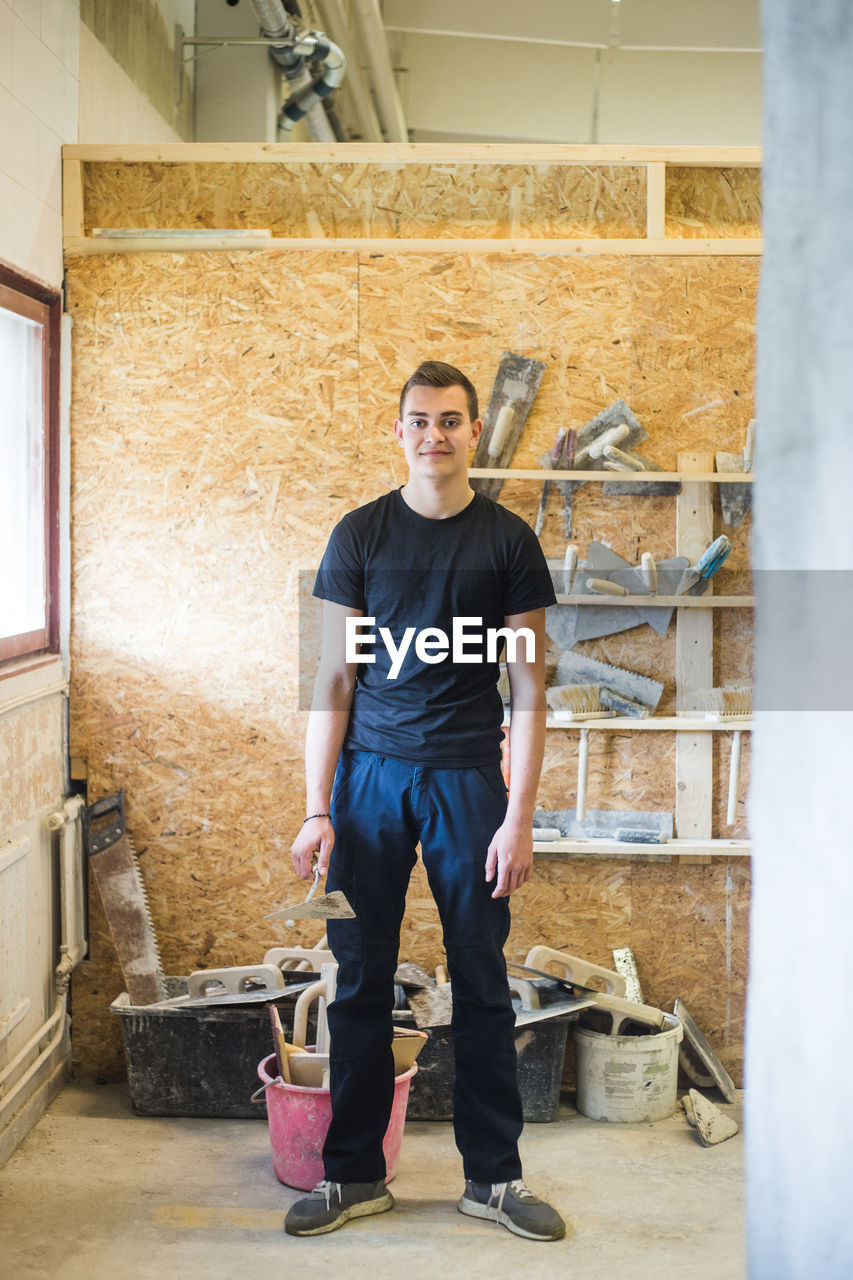 Full length portrait of confident young male trainee standing with equipment at workshop