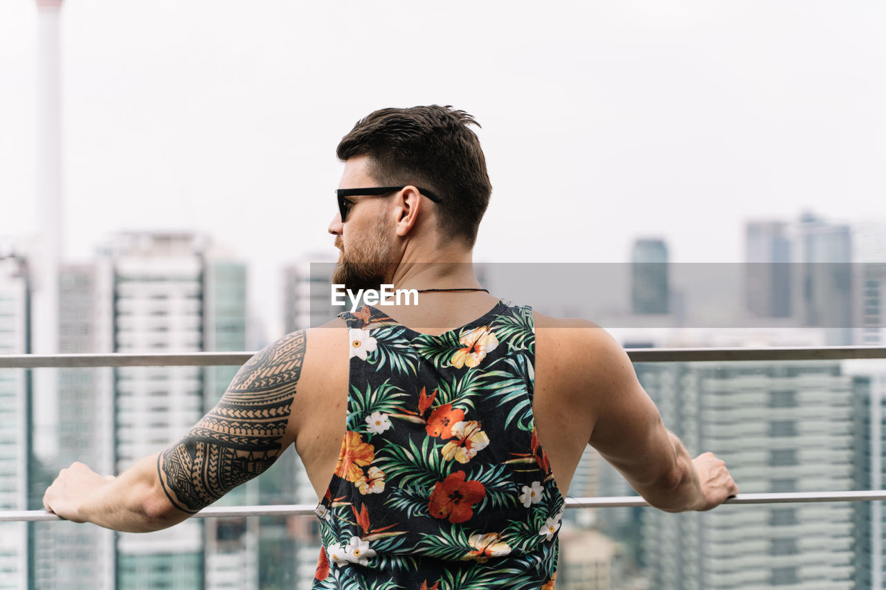 YOUNG MAN STANDING AGAINST MODERN CITY IN BACKGROUND