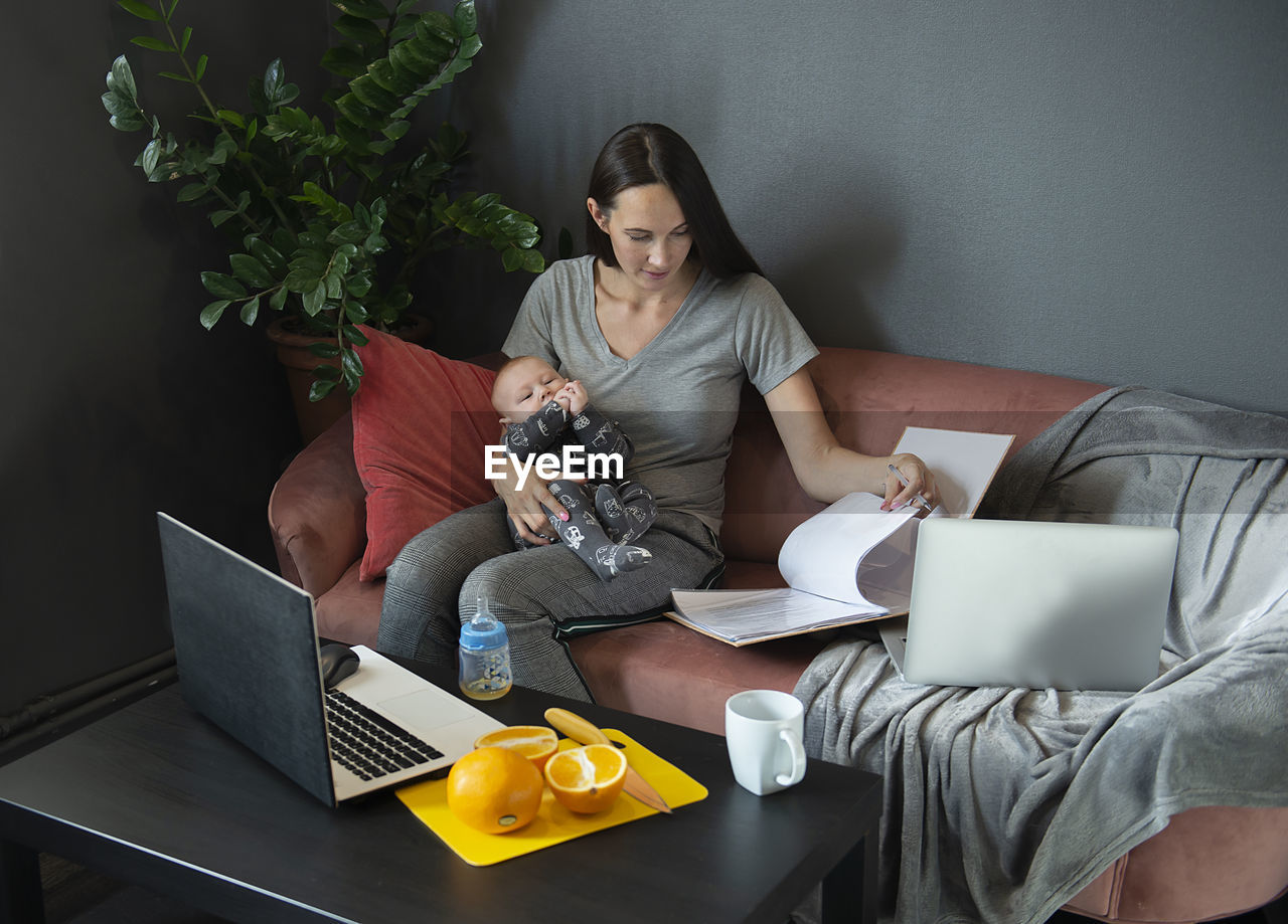 A young mom holds a baby and works from home with documents and laptops