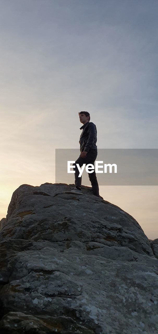 Low angle view of teenage boy standing on rock against sky