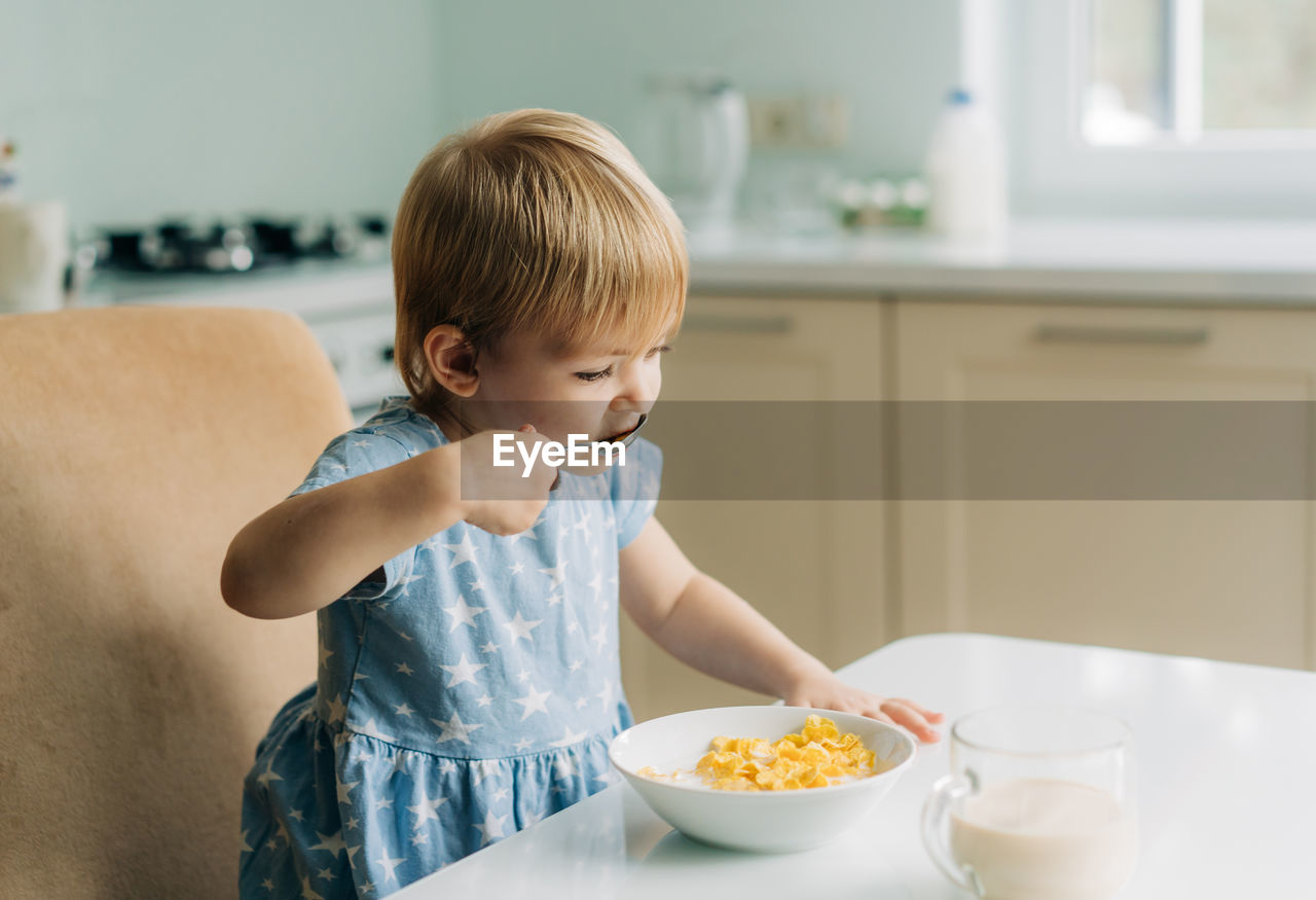 Little toddler girl eats corn flakes with milk in the kitchen in the morning.