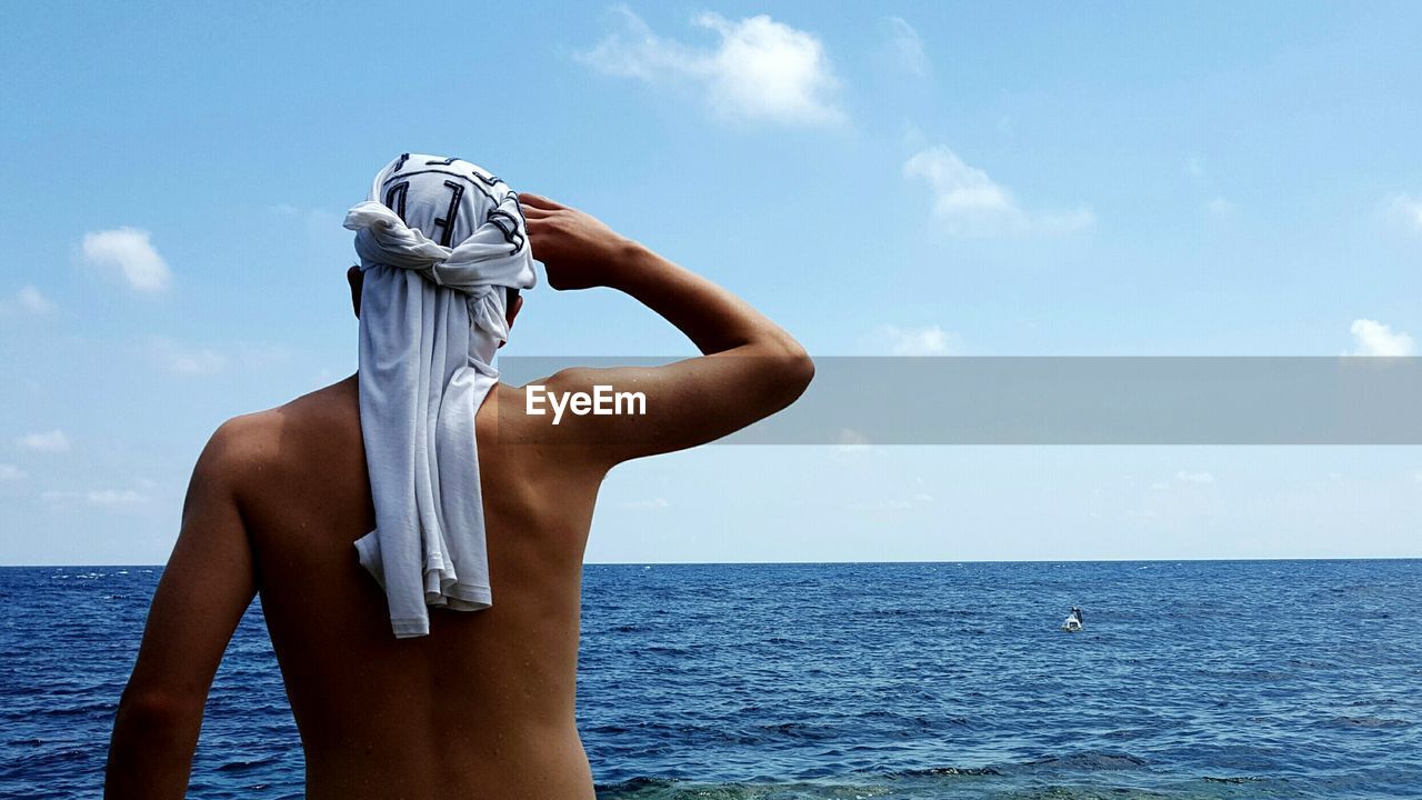 Rear view of shirtless man shielding eyes against sea
