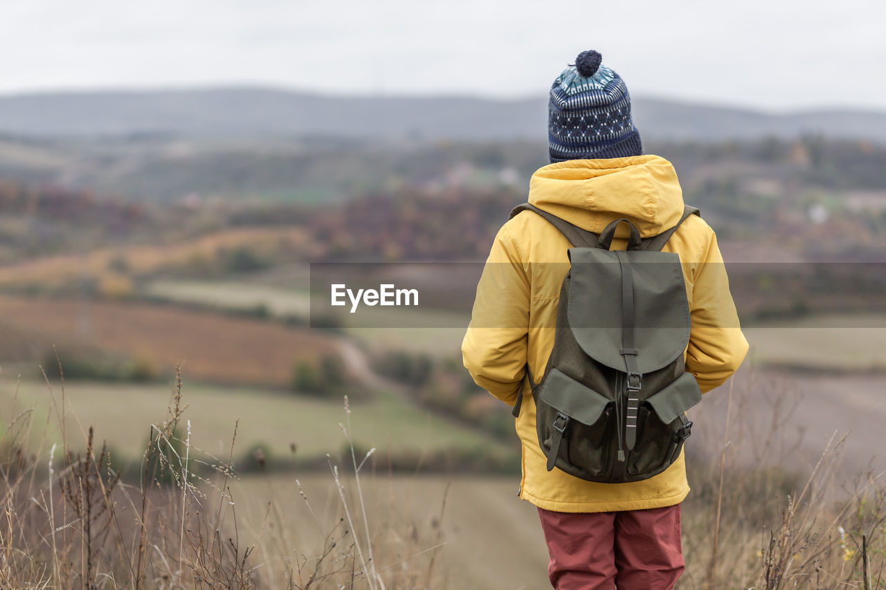 Rear view of a small boy with a backpack standing on a hill in autumn day and looking at view.