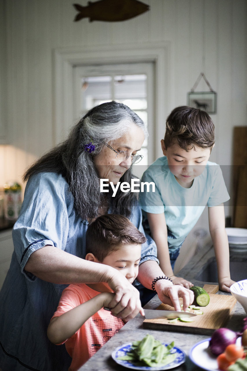 Grandmother assisting boy cutting cucumber on board in kitchen at home