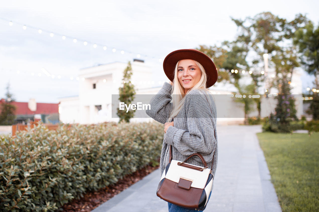 Smiling beautiful blonde teenage girl wear stylish hat, leather bag and knit gray jumper
