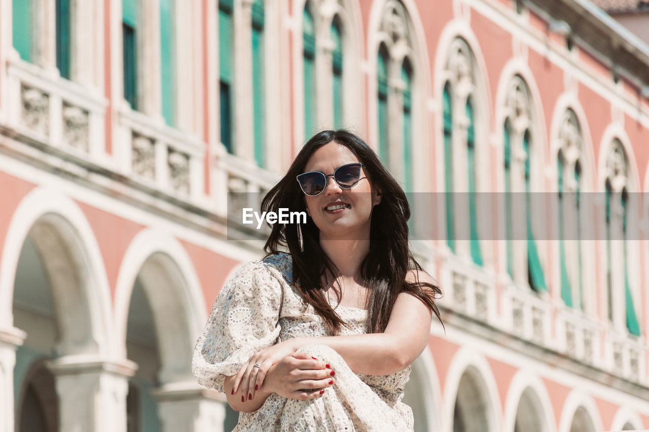 Portrait of beautiful young woman with long dark hair, sitting in square with beautiful architecture