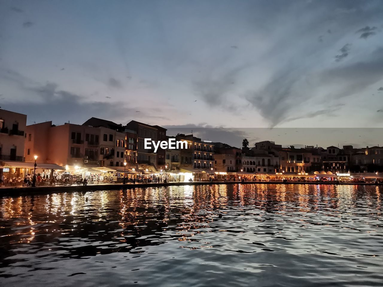 Buildings by illuminated city against sky at sunset