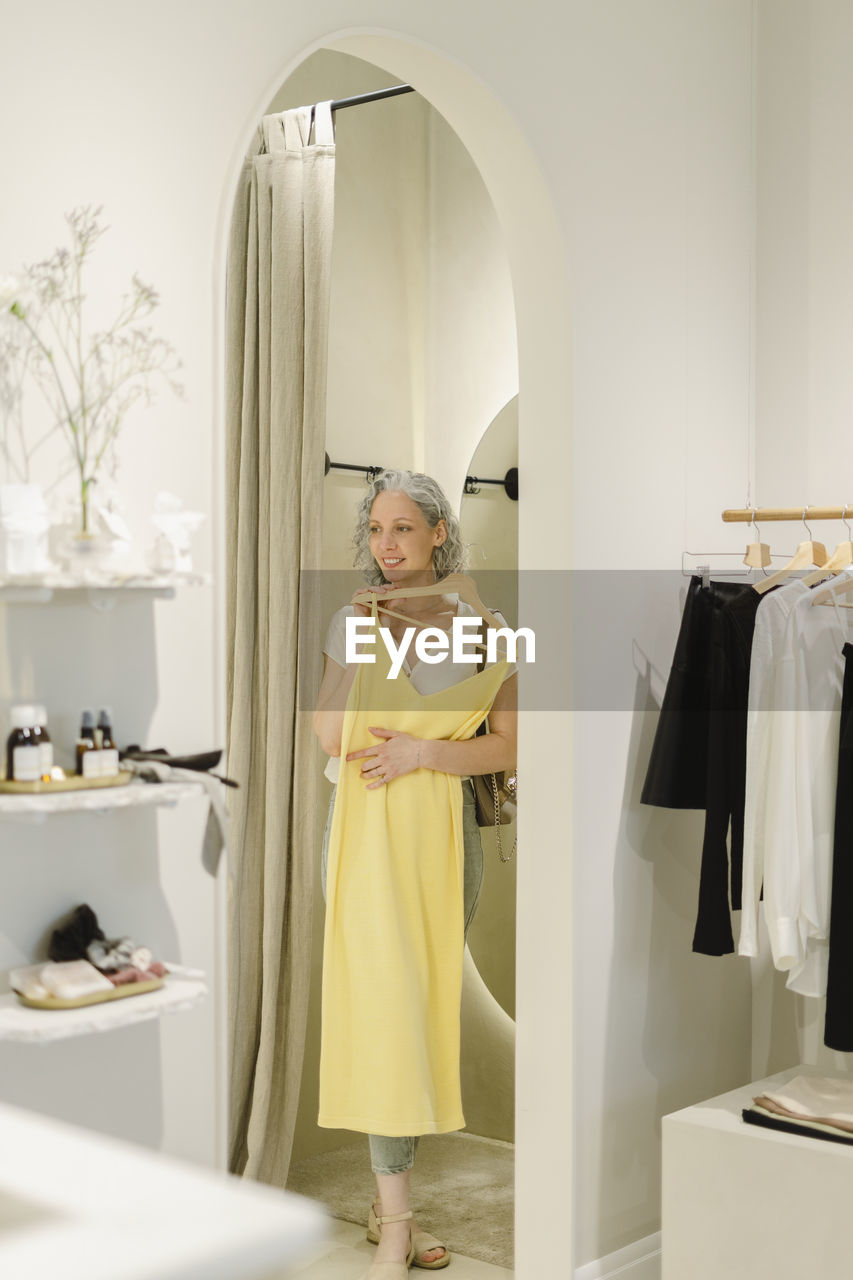 Smiling woman holding yellow dress stepping out from dressing room at store
