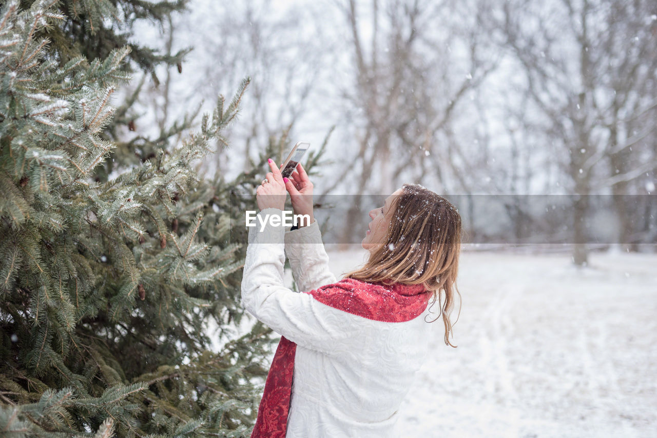 Woman photographing christmas tree during snowfall