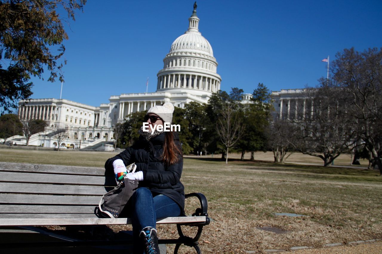 Young woman sitting on bench at park against state capitol building