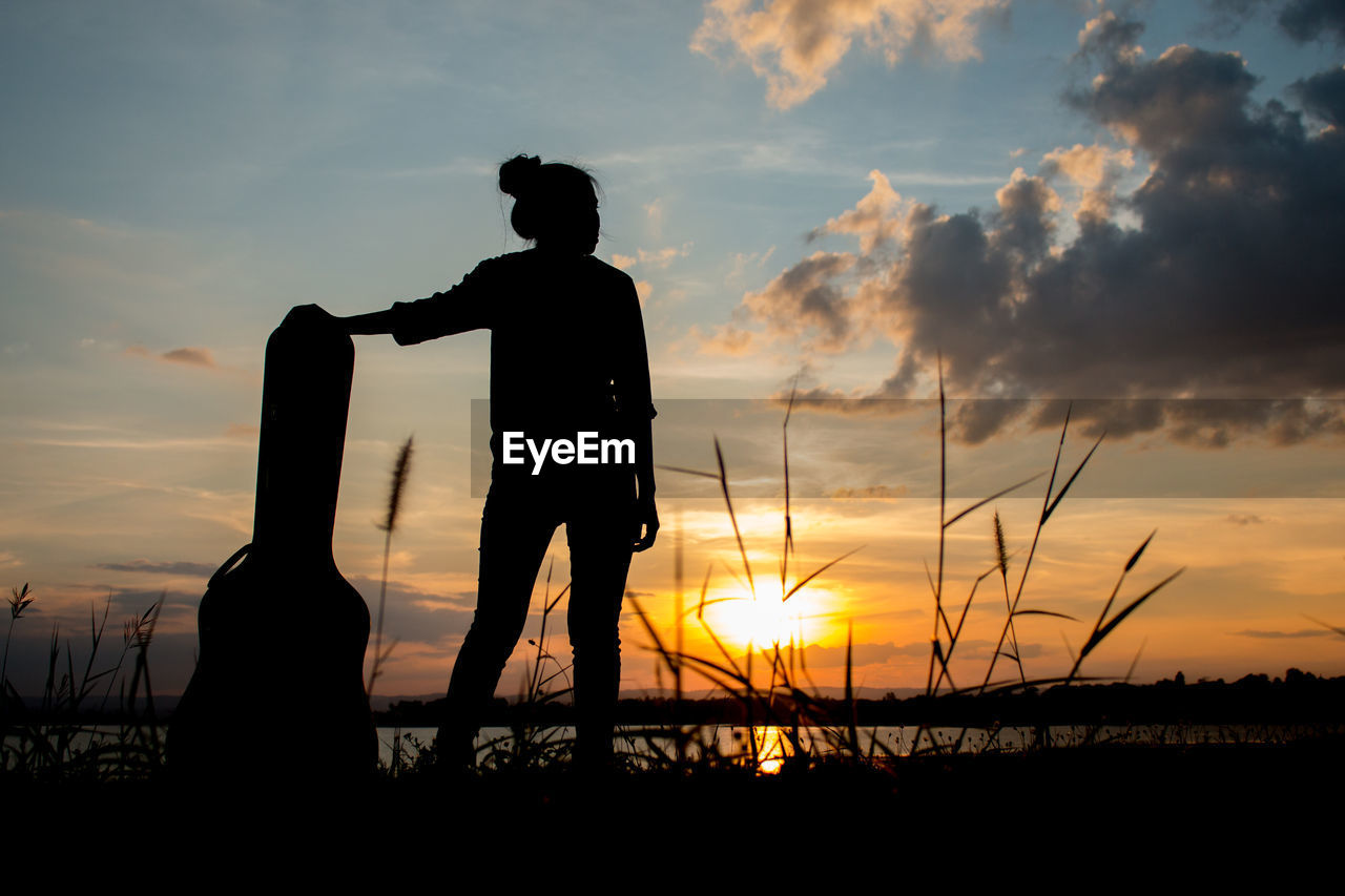 Silhouette woman standing with guitar case against sky during sunset