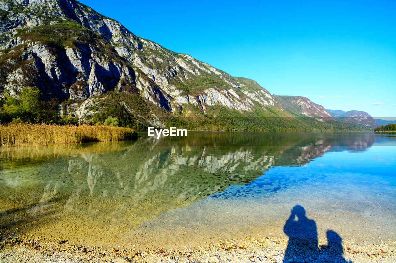 REAR VIEW OF MAN LOOKING AT LAKE AGAINST MOUNTAINS