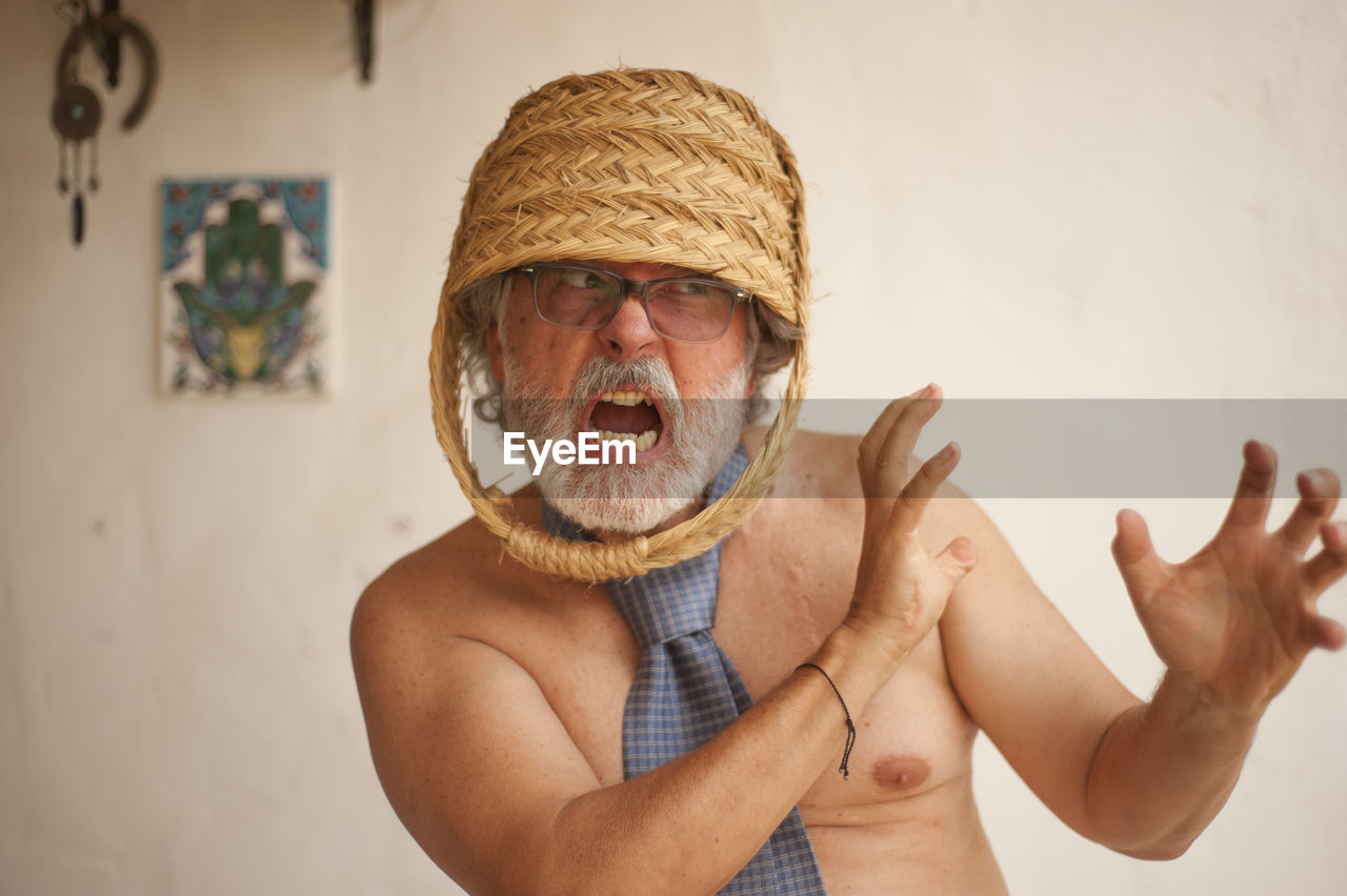 Angry senior man with basket on head standing against wall at home