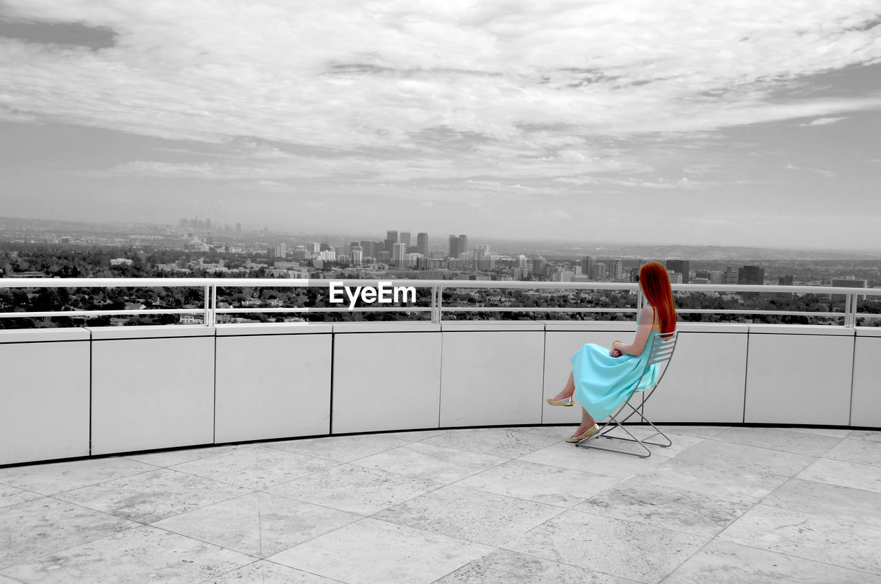 Woman looking at city while sitting on chair in building terrace against sky