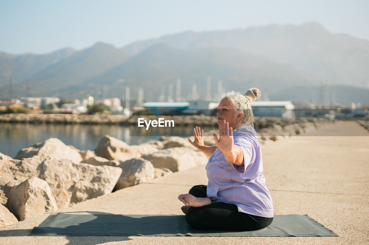 side view of woman sitting on rock by sea against mountain