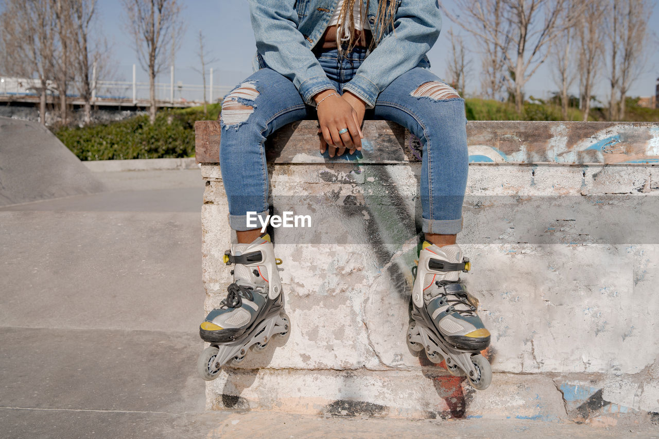 Crop black female with braided hairstyle and in rollerblades sitting on ramp in skate park and looking away
