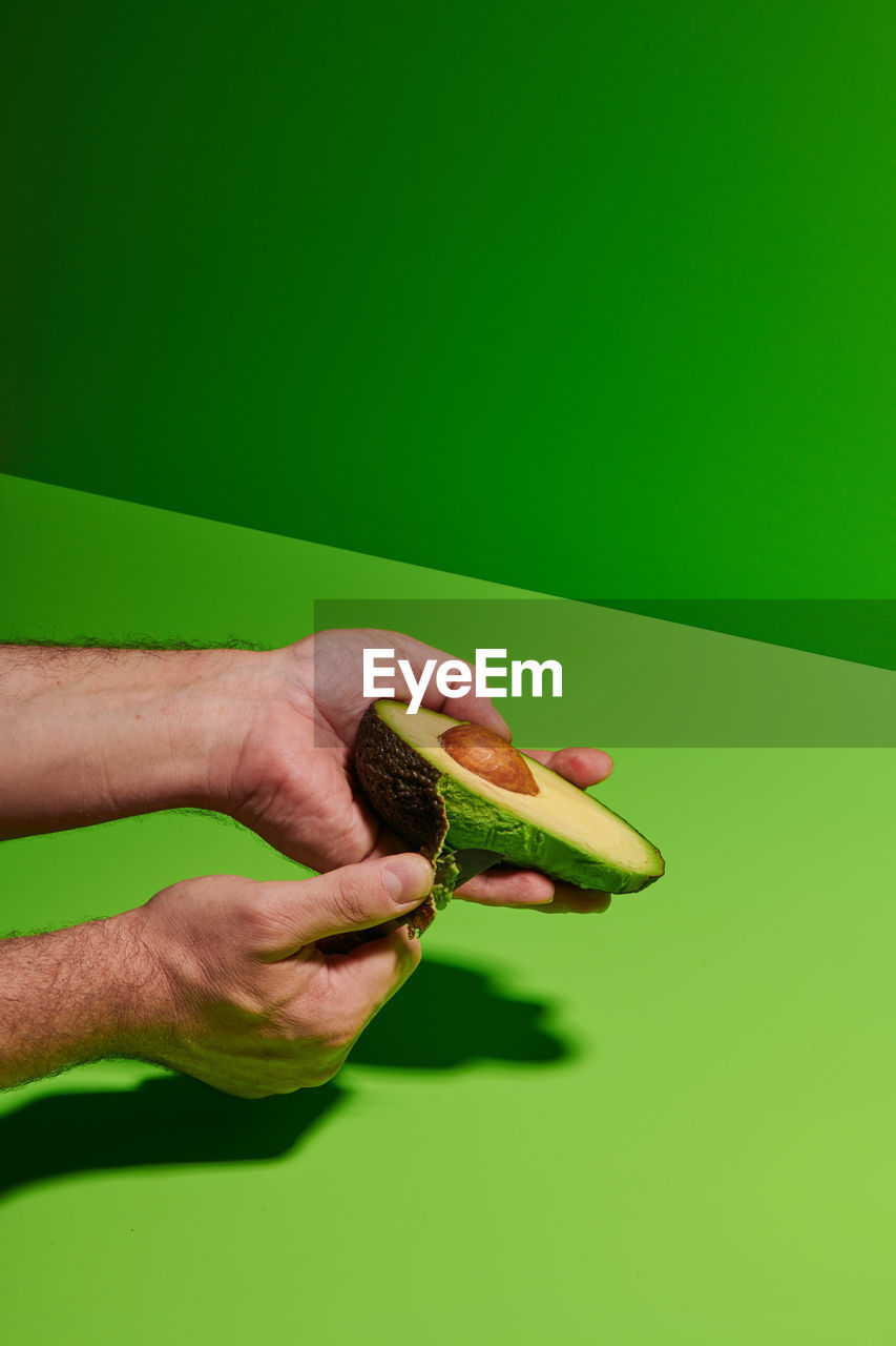 Crop unrecognizable person removing peel from fresh ripe avocado against bright green background