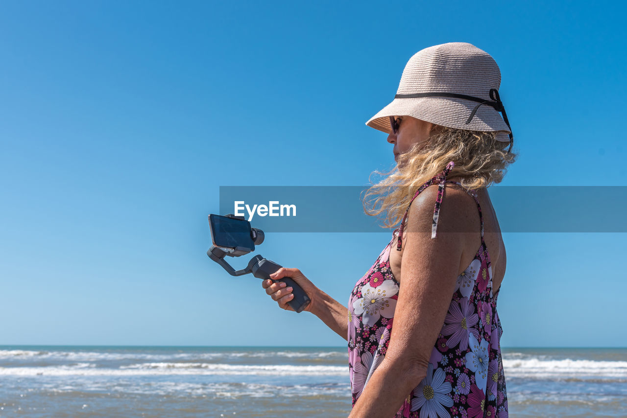 Blonde woman with a hat taking a selfie on the beach using a mobile attached to a gimbal