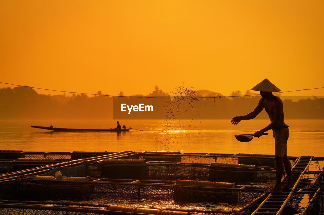 Silhouette fisherman feeding against orange sky