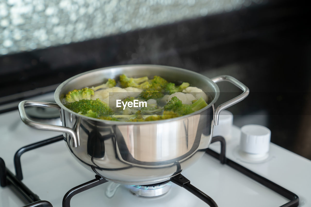 Green fresh broccoli is cooked in water in metal silver saucepan on gas white stove. 