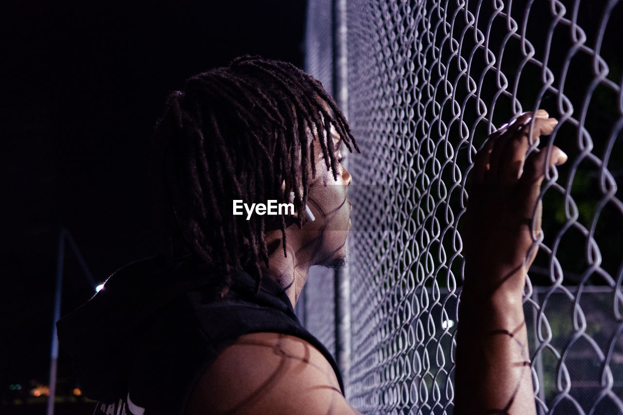 Side view of young man looking through chainlink fence at night