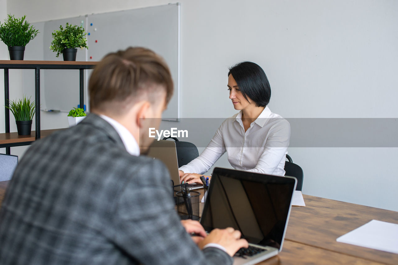 A man in a jacket is working at a laptop next to a woman in the office