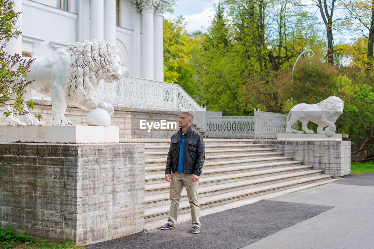 A man in casual clothes stands near a beautiful white building with lions