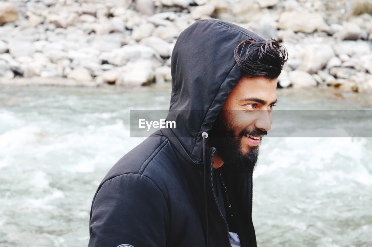 Smiling young man wearing hooded shirt while standing by river