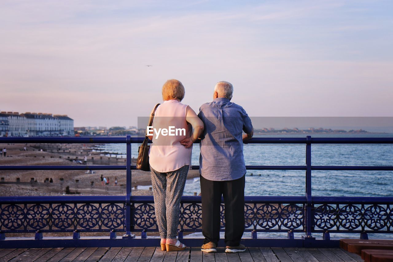 Rear view of couple looking at sea while standing by railing against sky during sunset