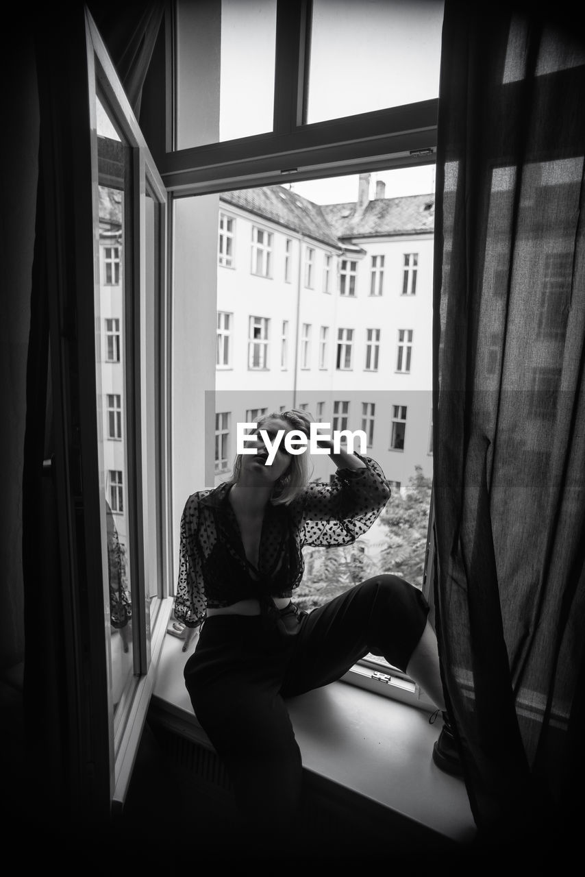 Full length of woman sitting on window sill at home